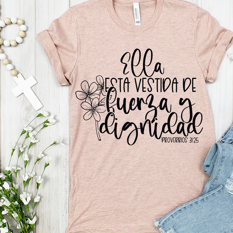 May include: A light pink t-shirt with rolled-up sleeves. The shirt features black script that reads "Ella Esta Vestida De Fuerza y Dignidad" with a floral design. The text is from Proverbs 31:25.