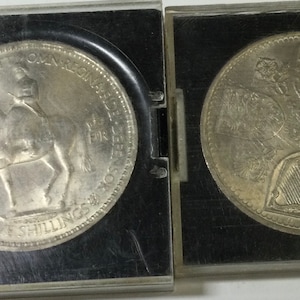 May include: Two silver coins in protective cases. The coin on the left features a profile of a person on horseback with the text "FID DEF GRATIA BRIT OMN REX FIDEI DEFENSOR" and "TWO SHILLINGS". The coin on the right features a design of four shields with the text "E II R" in the center.