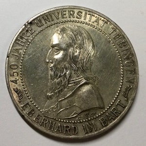 Könnte beinhalten: Eine silberne Münze mit einem detaillierten Profil eines Mannes mit Bart. Die Münze trägt die Inschrift "450 JAHRE UNIVERSITÄT TÜBINGEN EBERHARD IM BART" am Rand. Die Münze ist rund und hat einen detaillierten Rand.