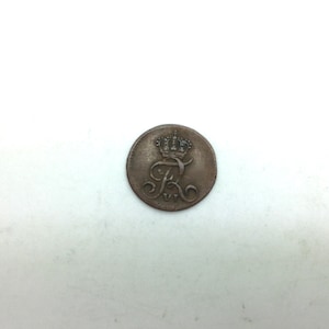 May include: A round, antique coin with a crown above the letter "R" and ornate scrollwork. The coin has a worn, aged appearance with a dark, metallic color. The design is centered on the coin, suggesting it may be a historical artifact or collectible.