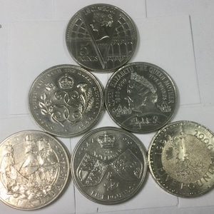 May include: Six silver coins with different designs. The coins feature the Queen Elizabeth II, a coat of arms, a ship, and a crown. The coins are all five pounds.