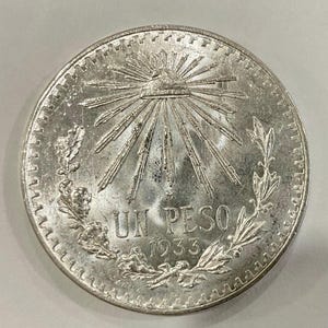 May include: A silver coin with the text "UN PESO" and the year "1933". The coin features a sunburst design with rays emanating from a central point, surrounded by a laurel wreath and other decorative elements.