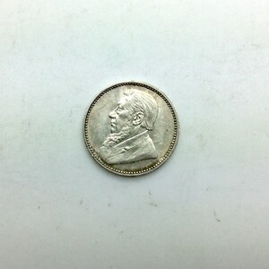 Puede incluir: Moneda de plata con un retrato de perfil de un hombre con barba y bigote. La moneda tiene un borde elevado y una superficie lisa.