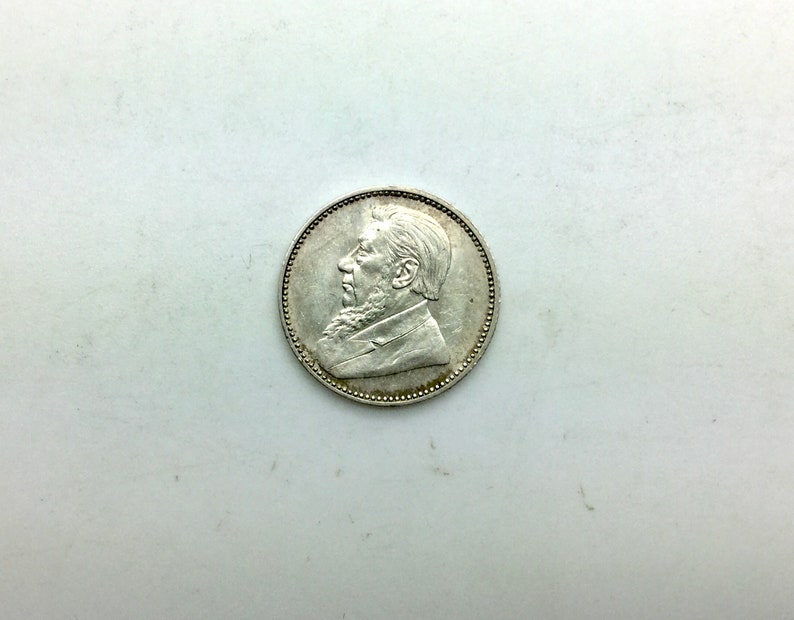 Puede incluir: Moneda de plata con un retrato de perfil de un hombre con barba y bigote. La moneda tiene un borde elevado y una superficie lisa.