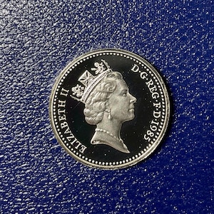 May include: A silver-colored coin featuring a profile portrait of Queen Elizabeth II wearing a crown and necklace. The text ELIZABETH II is on the left, and D.G.REG.F.D.1985 is on the right. The coin has a beaded border and a dark background.