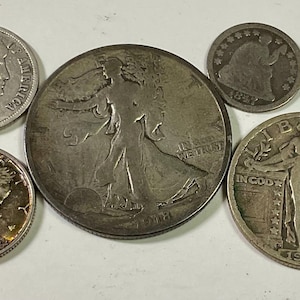 Op de afbeelding: Een verzameling antieke zilveren munten, waaronder een 1914 dime, een 1918 Walking Liberty half dollar, een 1926 Standing Liberty quarter en een Mercury dime. De munten zijn gerangschikt op een licht oppervlak.