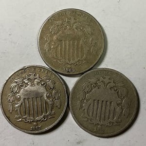 May include: Three antique nickel coins, each featuring a shield design with vertical stripes, surrounded by ornate detailing. The coins are dated 1867 and 1869, with the inscription "IN GOD WE TRUST" visible on each. The coins are silver in color.