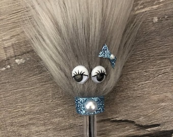 Op de afbeelding: Een grijze en witte pluche pentopper met oogjes, een blauwe glitterband, een parel en een blauwe strik met een parel accent.