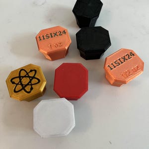 Puede incluir: Seis pequeños contenedores octogonales de varios colores, incluyendo rojo, blanco, negro, dorado y naranja. Los contenedores tienen el texto "11SIX24" y "1 oz" impreso en ellos.
