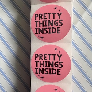 May include: A vertical roll of circular pink stickers featuring the black text PRETTY THINGS INSIDE in a bold sans-serif font. Each sticker is decorated with small black starburst icons positioned above and below the printed text.