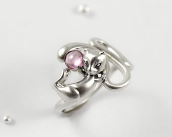 May include: A silver-toned ring shaped like a cat with a smooth, polished finish. The cat figure is depicted in a seated position, holding a small, round, translucent pink gemstone between its front paws. The band of the ring wraps around.