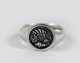 May include: A silver signet ring featuring a circular face with an engraved design of a crashing ocean wave and a mountain peak. The metal has a smooth, matte finish, and the recessed areas of the circular engraving are darkened to highlight the details.