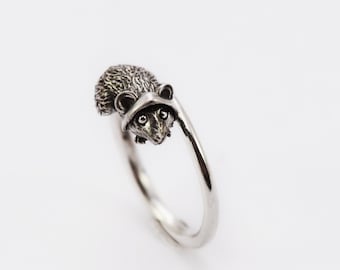May include: A silver-toned metal ring featuring a detailed, three-dimensional raccoon head and textured back. The band is smooth and polished, with the raccoon figure positioned at one end, creating an open-style design with a metallic finish.
