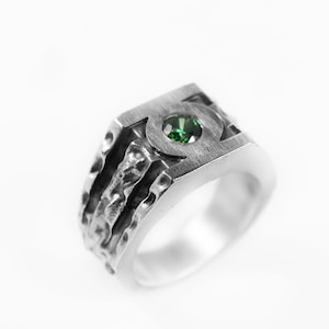 May include: A silver-toned ring featuring a brushed metal finish and a textured, uneven band design. The top of the ring displays a circular cutout containing a single, small, round-cut green gemstone set securely in the center of the metal surface.
