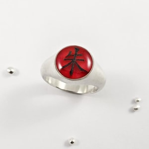 May include: A silver ring featuring a circular red inlay with a black Japanese kanji character centred on the face. The ring is surrounded by four small, loose silver beads scattered around the band on a plain white surface.