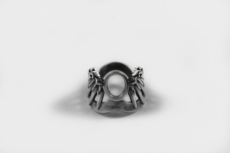 May include: A silver ring with a circular design featuring two wings with pointed tips. The wings are adorned with small, circular accents.