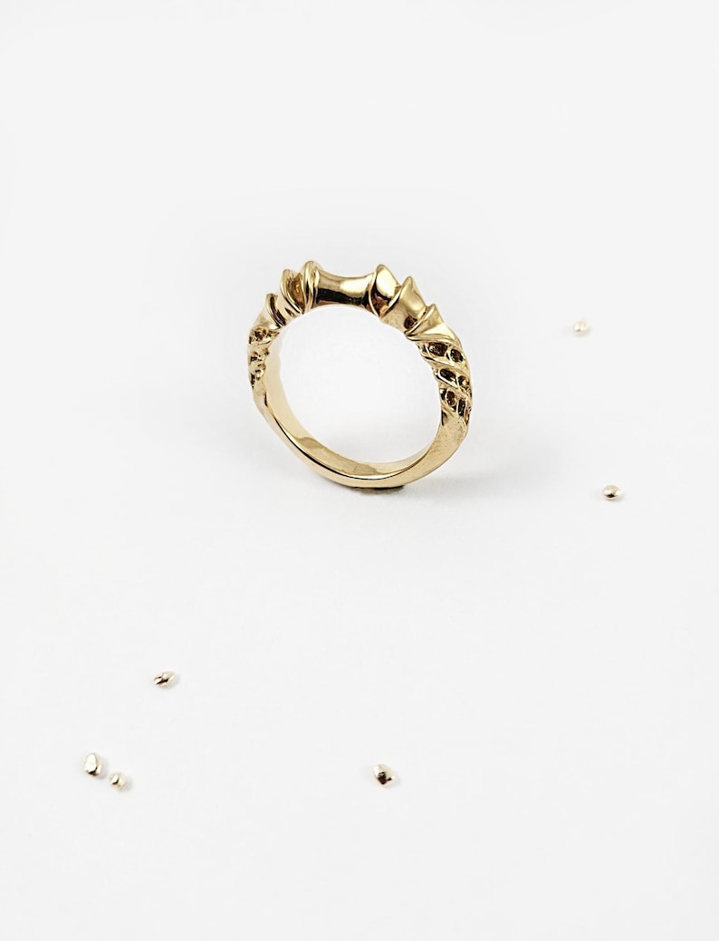May include: A gold ring featuring a textured, organic band with a series of raised, pointed elements along the top edge. The surface of the metal displays a detailed, uneven finish with small, scattered gold-toned beads nearby on the surface.