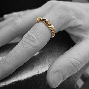 May include: A gold ring featuring a textured, intricate band with a repeating pattern of raised, organic shapes. The ring has a polished, metallic finish and is worn on a finger, standing out against the black and white background of the hand.