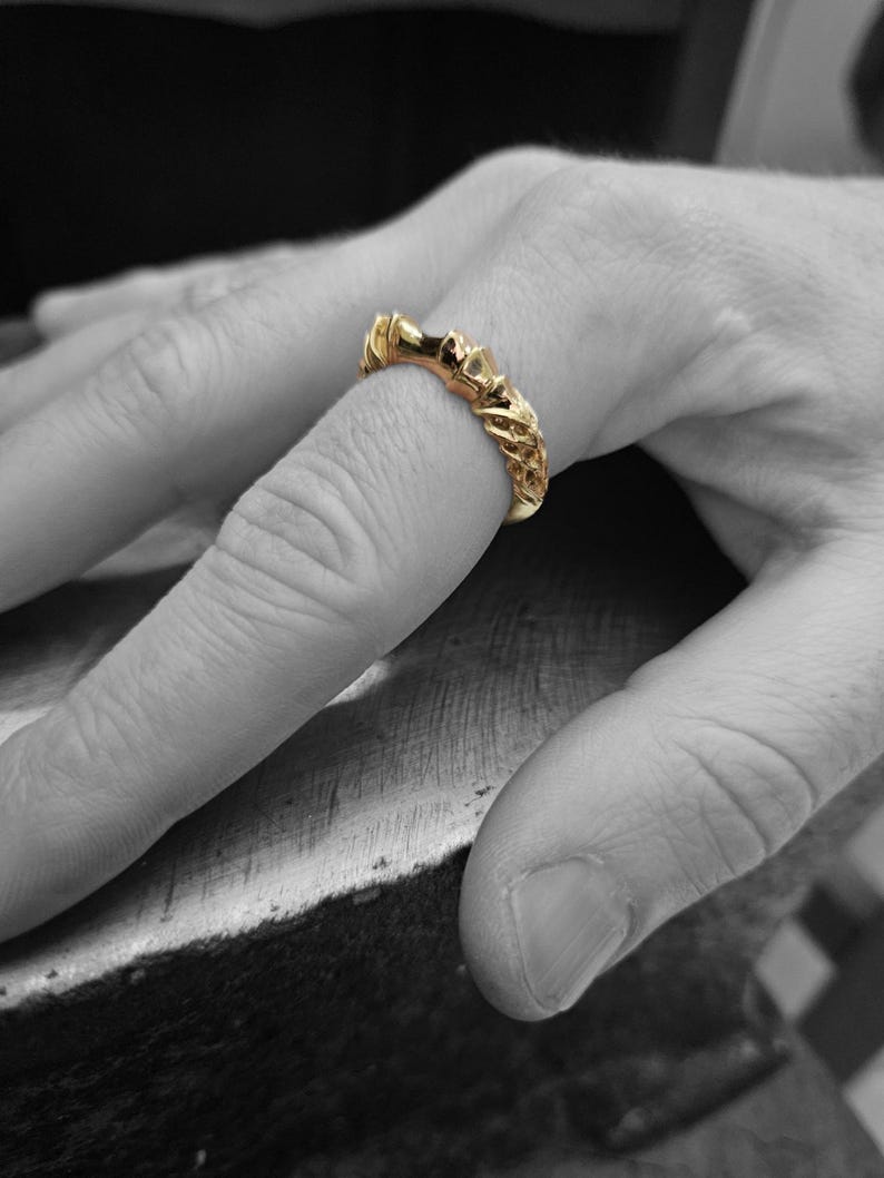 May include: A gold ring featuring a textured, intricate band with a repeating pattern of raised, organic shapes. The ring has a polished, metallic finish and is worn on a finger, standing out against the black and white background of the hand.