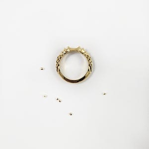 May include: A gold ring with a polished finish and a textured, woven-like pattern along the sides of the band. The ring features a slightly elevated, pointed design at the top center and is surrounded by several small, loose gold beads.