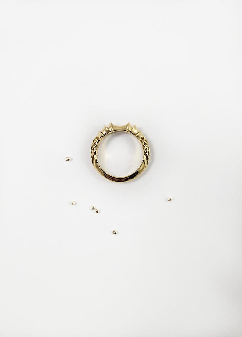May include: A gold ring with a polished finish and a textured, woven-like pattern along the sides of the band. The ring features a slightly elevated, pointed design at the top center and is surrounded by several small, loose gold beads.