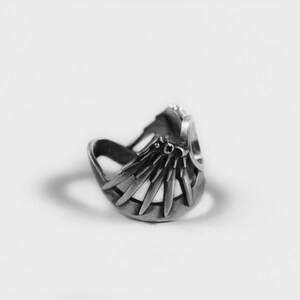 May include: A silver ring with a detailed design of a fan or a sunburst. The ring is open on one side and has a curved shape.