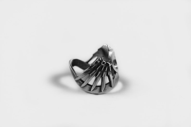 May include: A silver ring with a detailed design of a fan or a sunburst. The ring is open on one side and has a curved shape.