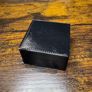 May include: A black, square-shaped box with a flat lid and smooth vertical sides. The top surface of the lid features a visible diagonal cross-hatch texture pattern, while the sides exhibit horizontal layer lines consistent with 3D printing.