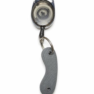 May include: A retractable badge reel featuring a circular, translucent gray housing with a silver-toned carabiner clip at the top. A black cord extends from the reel to a silver metal split ring, which holds a textured, curved gray plastic tag.