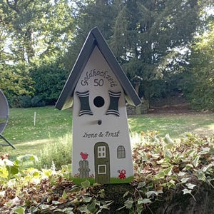 May include: A white birdhouse with a gray roof and decorative accents. The birdhouse has the text "Goldhochzeit 50" and the names "Irene & Ernst". A red bird and mushroom are painted on the front. The birdhouse is sitting on a stone wall.