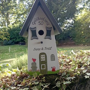 May include: A white birdhouse with a gray roof and decorative accents. The birdhouse features the text "goldhochzeit 50" and the names "Irene & Ernst". A painted bird, door, and window are also visible.