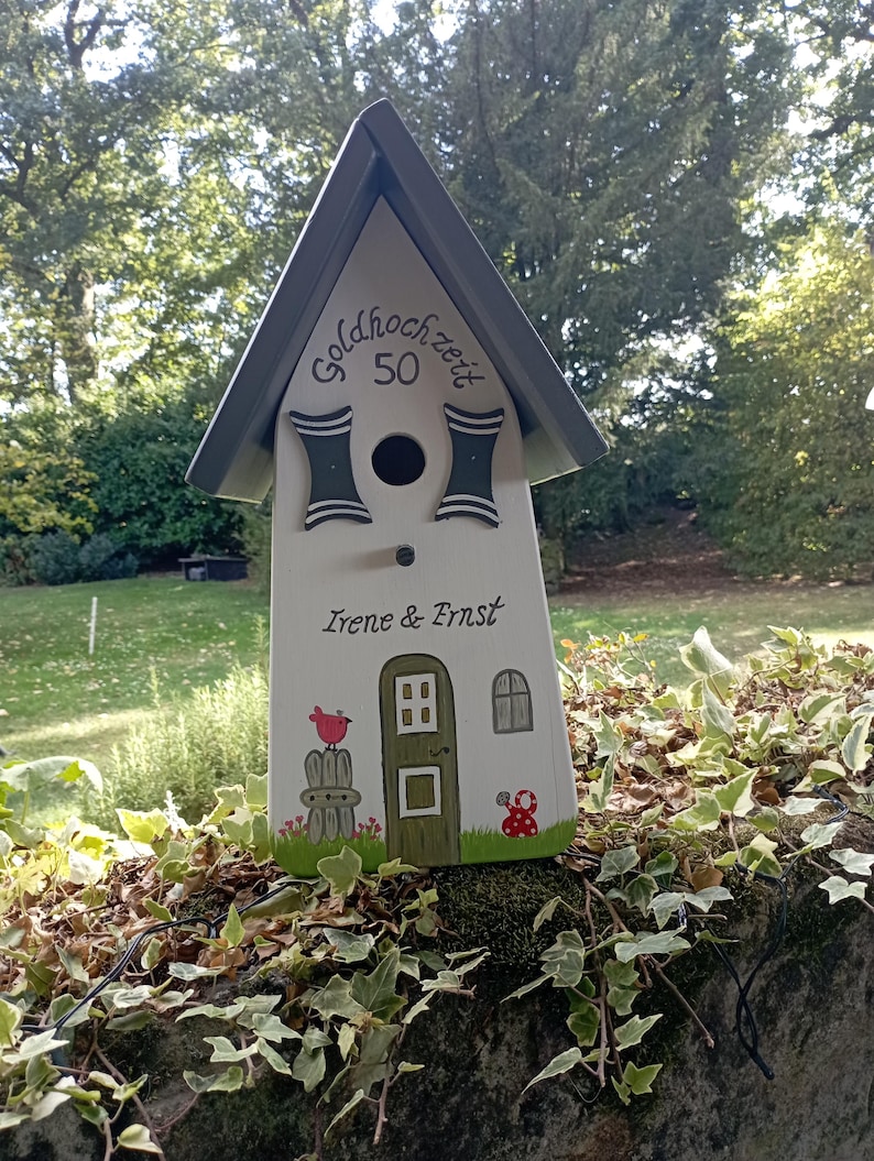 May include: A white birdhouse with a gray roof and decorative accents. The birdhouse features the text "goldhochzeit 50" and the names "Irene & Ernst". A painted bird, door, and window are also visible.