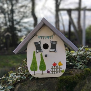 May include: A decorative white birdhouse with a gray roof, featuring painted green trees, a fence with birds, and a banner. The birdhouse has two black and white window designs and a round entry hole. It is sitting on a mossy rock.