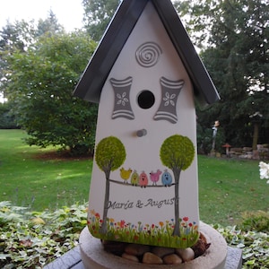 Puede incluir: Una casita para p&aacute;jaros blanca con techo gris y detalles decorativos. La casita tiene un agujero de entrada circular y presenta un dise&ntilde;o pintado de &aacute;rboles, p&aacute;jaros de colores y el texto "Maria & August". Se encuentra sobre una jardinera de hormig&oacute;n llena de piedras.