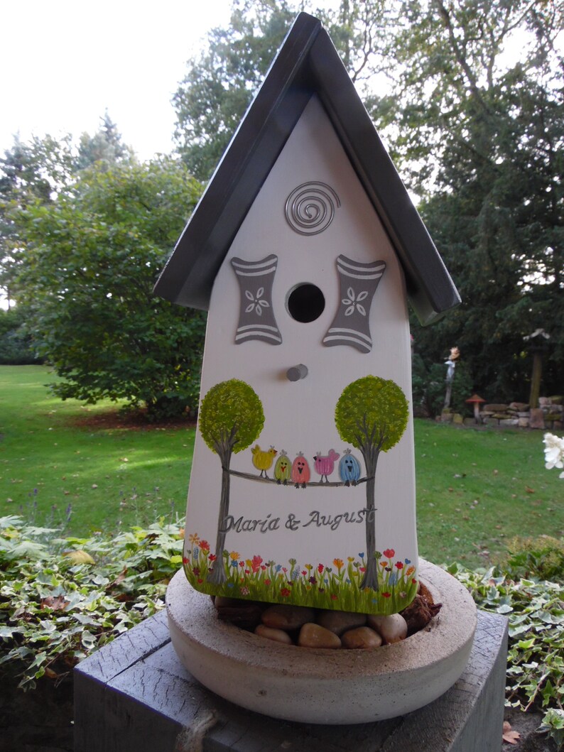 Puede incluir: Una casita para p&aacute;jaros blanca con techo gris y detalles decorativos. La casita tiene un agujero de entrada circular y presenta un dise&ntilde;o pintado de &aacute;rboles, p&aacute;jaros de colores y el texto "Maria & August". Se encuentra sobre una jardinera de hormig&oacute;n llena de piedras.