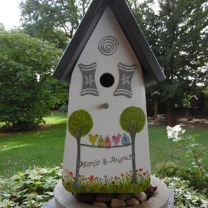 Puede incluir: Una pajarera blanca con techo gris y detalles decorativos. La pajarera presenta un dise&ntilde;o pintado de &aacute;rboles, p&aacute;jaros y el texto "Maria & August". Se encuentra sobre una jardinera de hormig&oacute;n llena de piedras.