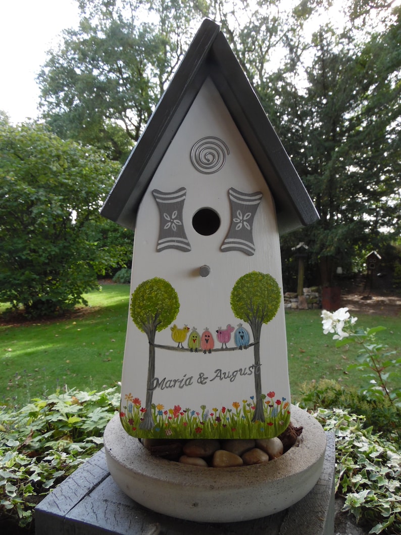 Puede incluir: Una pajarera blanca con techo gris y detalles decorativos. La pajarera presenta un dise&ntilde;o pintado de &aacute;rboles, p&aacute;jaros y el texto "Maria & August". Se encuentra sobre una jardinera de hormig&oacute;n llena de piedras.