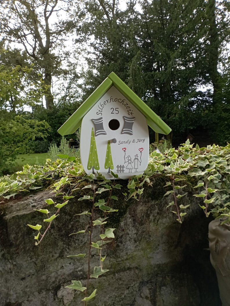May include: A white birdhouse with a green roof and trim, featuring the text "Silberhochzeit 25" and the names "Sandy & Jorg." The birdhouse has a round entry hole and is decorated with green tree and couple illustrations. It rests on a stone wall.