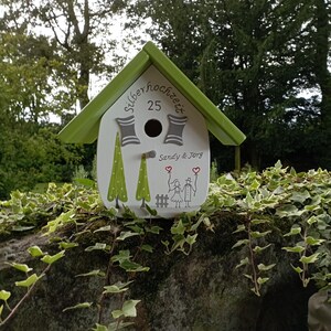 May include: A white birdhouse with a green roof, featuring the text "Silberhochzeit 25" and the names "Sandy & Jorg." The birdhouse is decorated with green trees and a drawing of two people holding hands. The birdhouse is sitting on a stone wall.