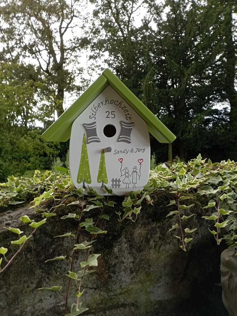 May include: A white birdhouse with a green roof, featuring the text "Silberhochzeit 25" and the names "Sandy & Jorg." The birdhouse is decorated with green trees and a drawing of two people holding hands. The birdhouse is sitting on a stone wall.