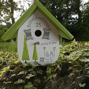 May include: A decorative birdhouse with a green roof and white body. The birdhouse features the text "silberhochzeit 25" and the names "Sandy & J&ouml;rg". It is decorated with painted trees and a couple holding hearts.