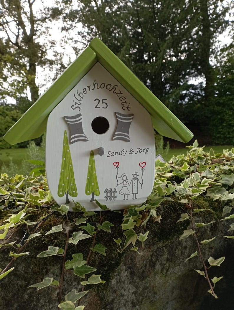 May include: A decorative birdhouse with a green roof and white body. The birdhouse features the text "silberhochzeit 25" and the names "Sandy & J&ouml;rg". It is decorated with painted trees and a couple holding hearts.