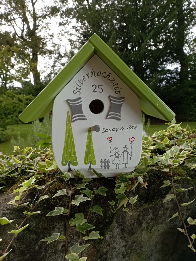 May include: A decorative birdhouse with a green roof and white body. The birdhouse features the text "silberhochzeit 25" and the names "Sandy & J&ouml;rg". It is decorated with painted green trees and a couple holding hearts.