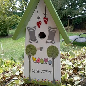 Może przedstawiać: Biały drewniany domek dla ptak&oacute;w z zielonym dachem i dekoracyjnymi malowanymi detalami. Domek dla ptak&oacute;w przedstawia rząd kolorowych ptak&oacute;w siedzących na drucie, drzewa i napis "Villa L&ouml;ber". Czerwone ozdoby w kształcie serca wiszą z dachu.