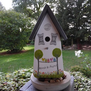 Puede incluir: Una casita para p&aacute;jaros blanca con techo gris y detalles decorativos. La casita presenta &aacute;rboles pintados, p&aacute;jaros coloridos y los nombres "Maria & August". Se encuentra en una jardinera de hormig&oacute;n llena de piedras, con c&eacute;sped verde y &aacute;rboles al fondo.