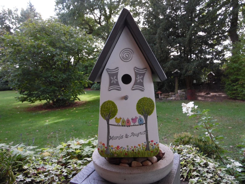 Puede incluir: Una casita para p&aacute;jaros blanca con techo gris y detalles decorativos. La casita presenta &aacute;rboles pintados, p&aacute;jaros coloridos y los nombres "Maria & August". Se encuentra en una jardinera de hormig&oacute;n llena de piedras, con c&eacute;sped verde y &aacute;rboles al fondo.