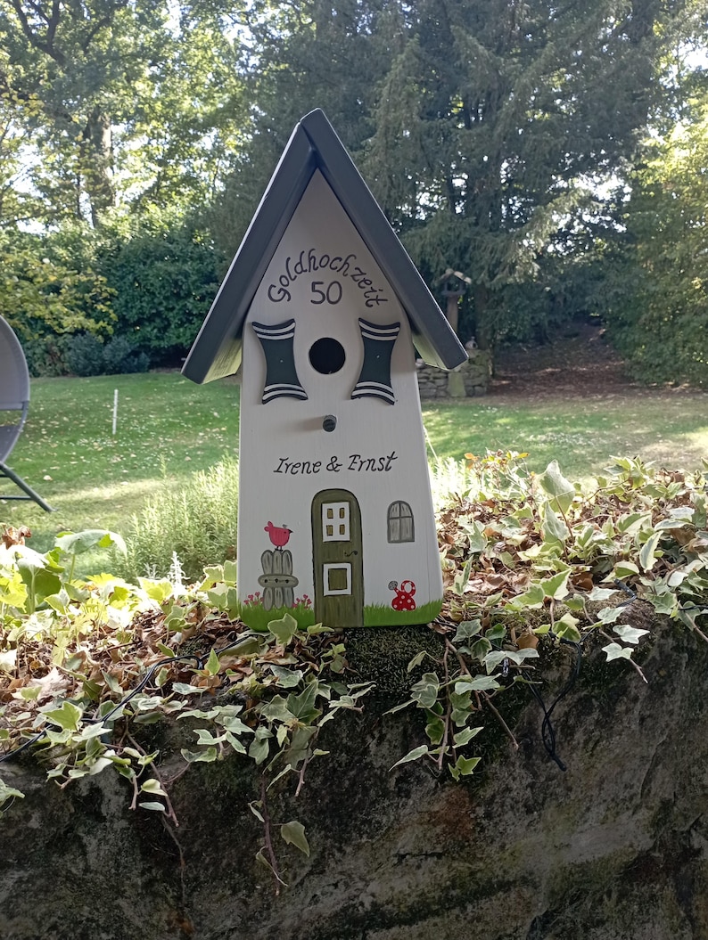 May include: A white birdhouse with a gray roof and decorative accents. The birdhouse has the text "Goldhochzeit 50" and the names "Irene & Ernst". A red bird and mushroom are painted on the front. The birdhouse is sitting on a stone wall.