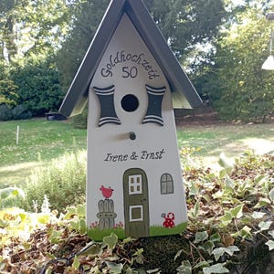 May include: A decorative birdhouse painted white with a gray roof. The birdhouse has a door, window, and a red bird perched on a fence. The text "Goldhochzeit 50" and "Irene & Ernst" are painted on the birdhouse.