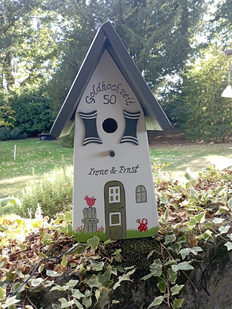 May include: A decorative birdhouse painted white with a gray roof. The birdhouse has a door, window, and a red bird perched on a fence. The text "Goldhochzeit 50" and "Irene & Ernst" are painted on the birdhouse.