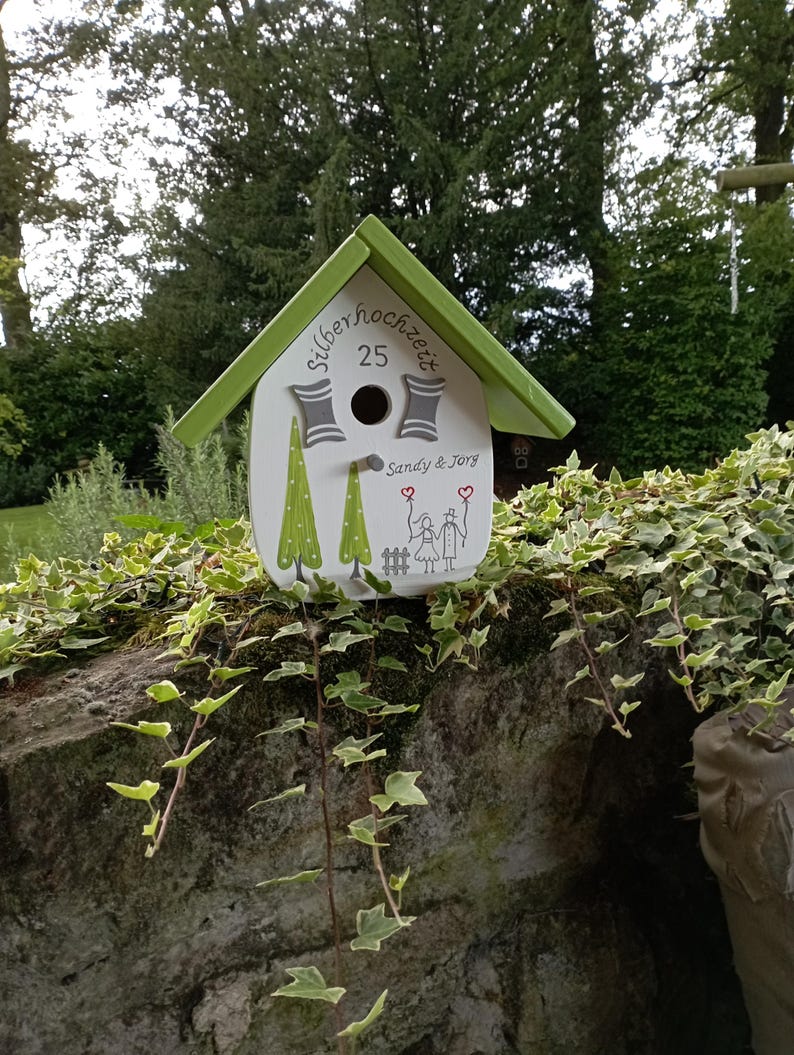 May include: A white birdhouse with a green roof and trim, decorated with painted trees and the text "silberhochzeit 25". The birdhouse has a round entry hole and a drawing of two stick figures holding hands.